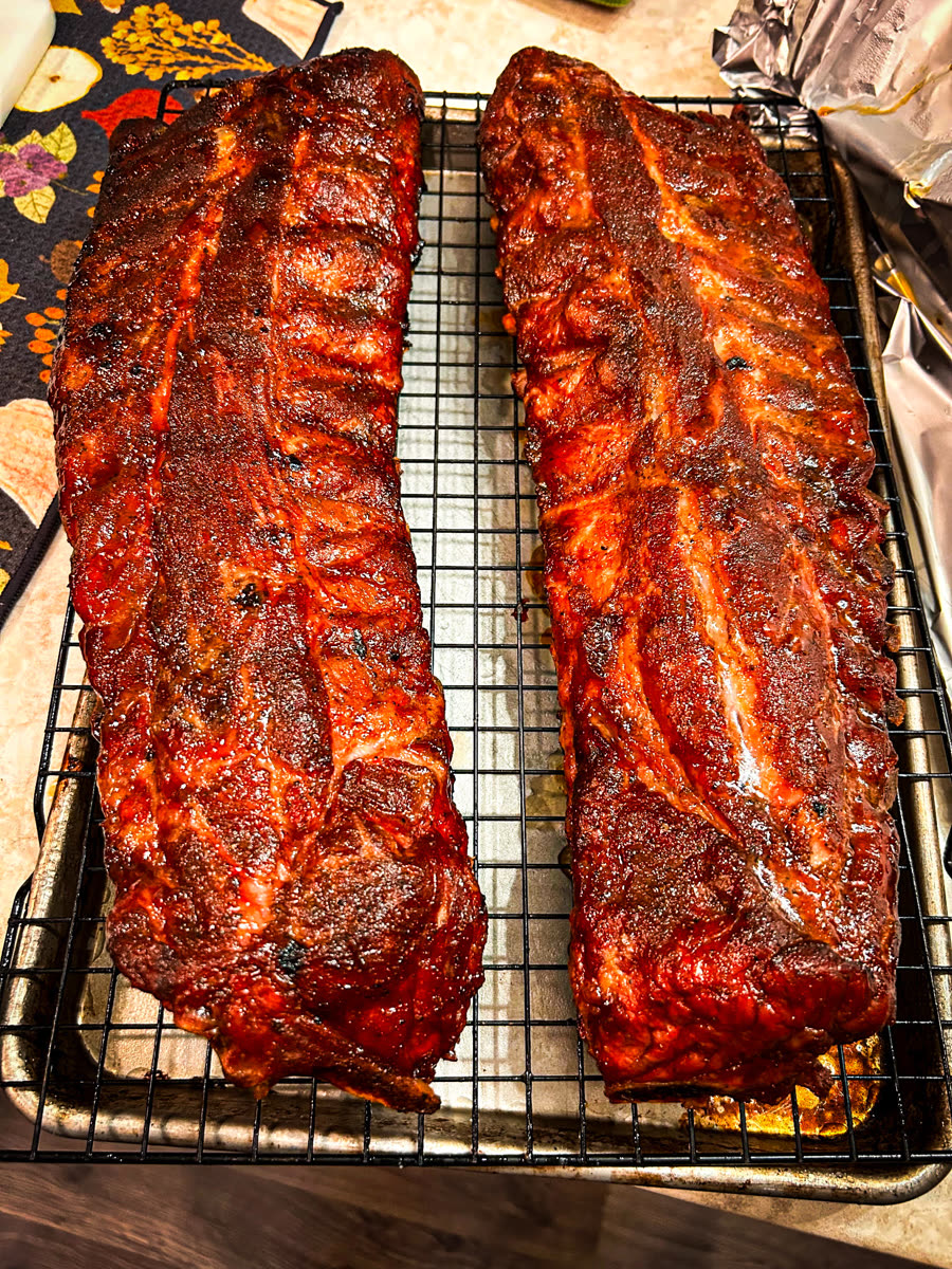 Two full racks of smoked pork ribs with a glossy, sauced bark resting on a wire rack