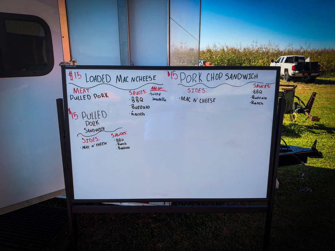 Whiteboard menu listing loaded mac and cheese, pulled pork sandwich, and pork chop sandwich, fifteen dollars each