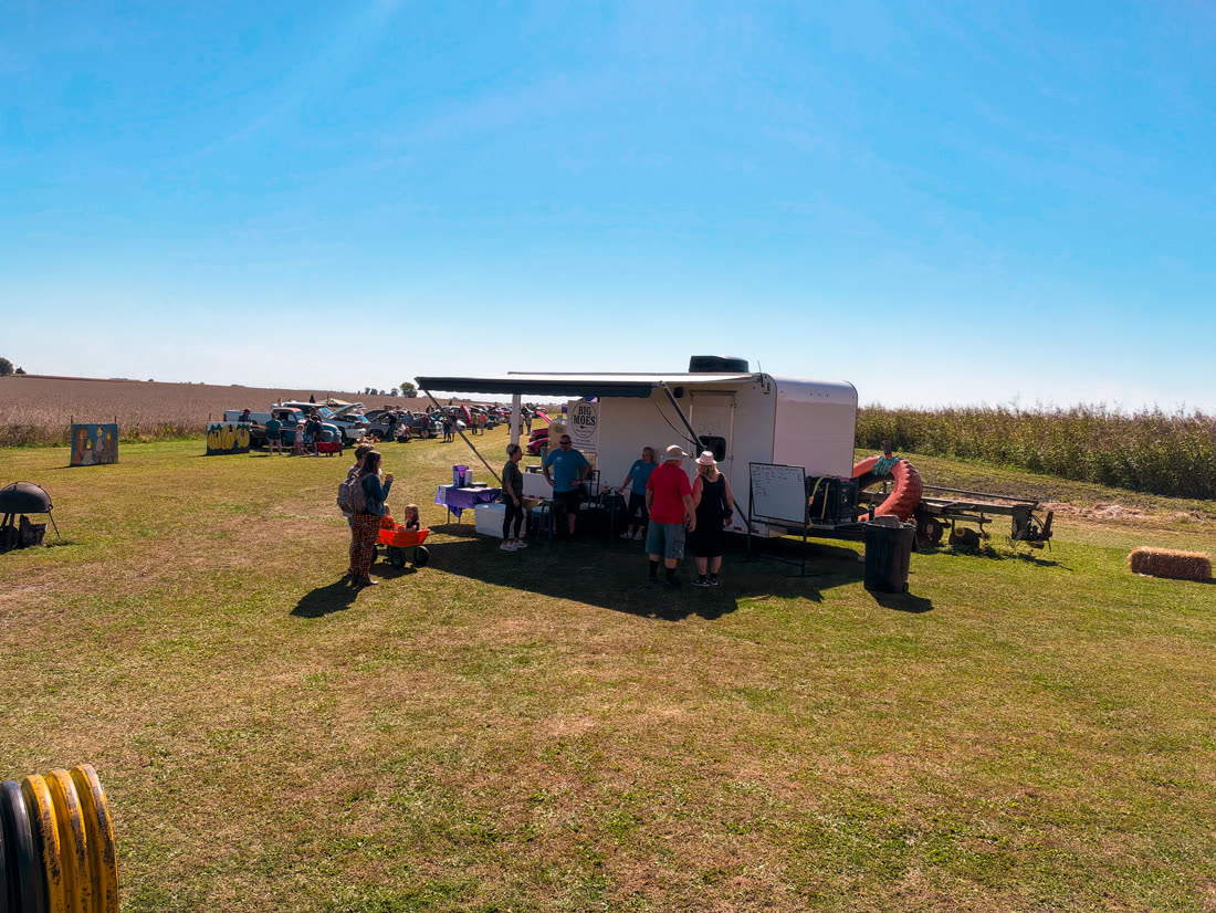 Wide view of the Big Moe's trailer set up on the grass next to the car show and cornfields